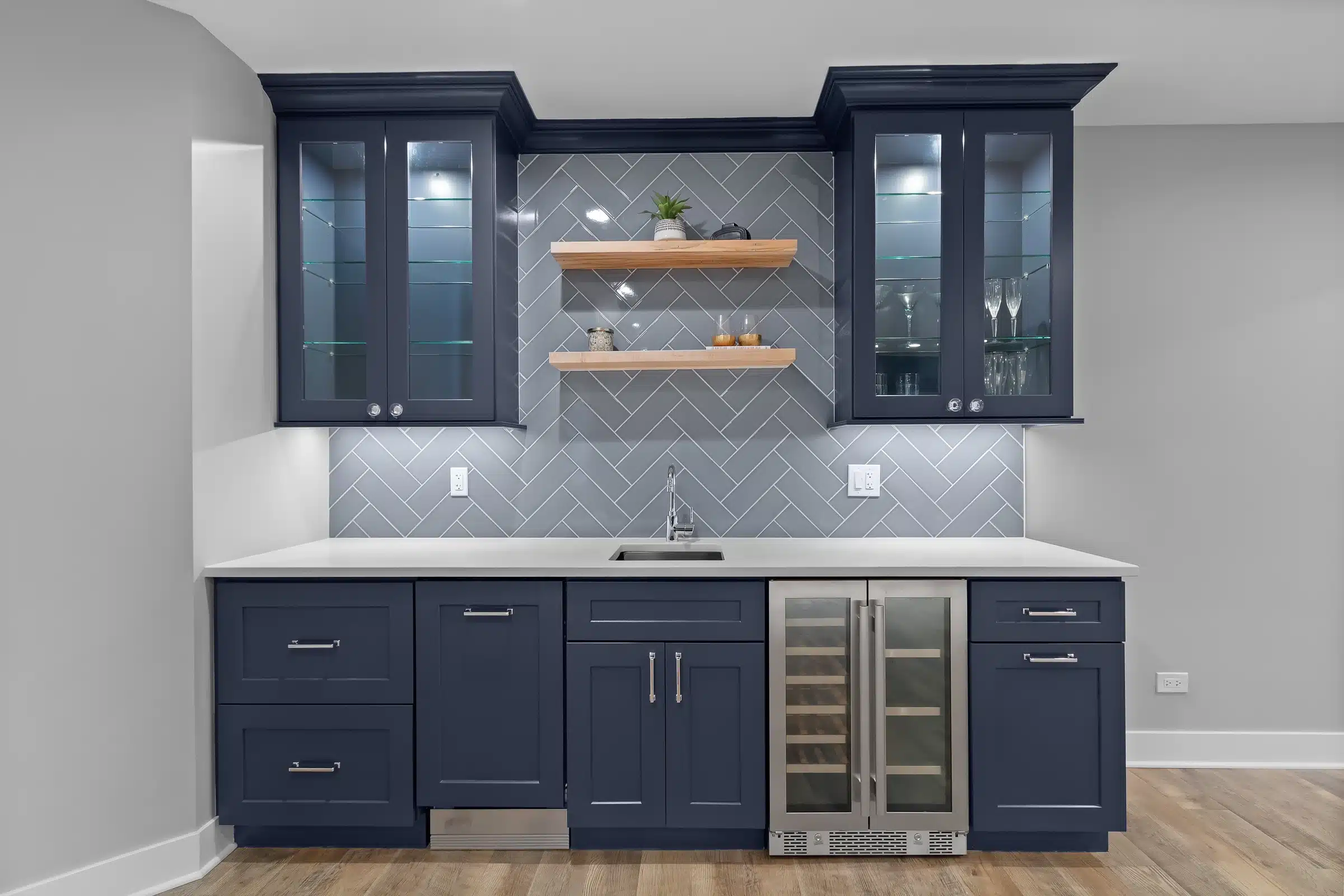 Close up of custom navy blue basement wet bar with gray herringbone backsplash, light wood floating shelves, and glass front upper cabinets in Palatine.