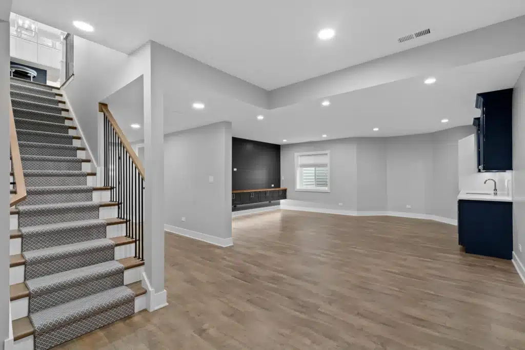 Wide angle view of an open concept finished basement renovation in Palatine, featuring luxury vinyl plank flooring, a carpeted staircase, a black shiplap media wall, and a navy blue wet bar area.