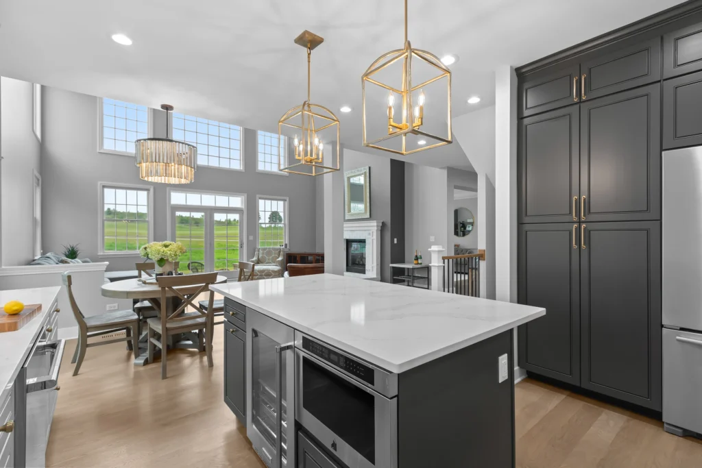 Wide angle view of an open concept kitchen in Palatine featuring a charcoal island, gold cage pendant lights, and a view into a sun-filled living room with vaulted ceilings.