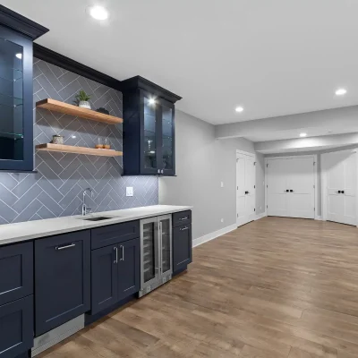Wide angle view of a finished basement in Palatine featuring a navy blue wet bar with quartz counters, opening onto luxury vinyl plank flooring and double interior doors.