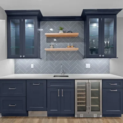 Close up of custom navy blue basement wet bar with gray herringbone backsplash, light wood floating shelves, and glass front upper cabinets in Palatine.